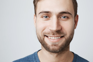 Obraz premium Close-up portrait of happy young male model with beard and stylish hairdo, smiles pleasantly and broadly with teeth at camera, expresses positive emotions, isolated against gray background