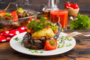 Eggplants stuffed with minced meat and cooked with fresh tomatoes and bell peppers on white plate and in baking dish on wooden rustic table.