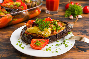 Eggplants stuffed with minced meat and cooked with fresh tomatoes and bell peppers on white plate and in baking dish on wooden rustic table.