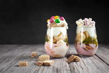 Dessert in a glass with a banana, cookies, candies, waffles and jelly on a wooden table