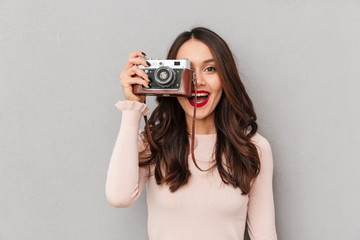 Image of pretty female photographer holding retro camera doing favorite shots isolated against gray...