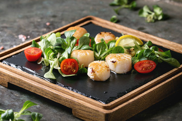 Fried scallops with lemon, cherry tomatoes and green salad served on wooden black slate serving...