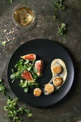 Fried scallops with lemon, figs, sauce and green salad served on black plate with glass of white wine over old dark metal background. Top view, space. Plating, fine dining - 188218147