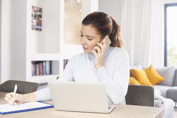 Managing my business from home. A beautiful middle aged woman using laptop and mobile phone while sitting at desk and working from home. Home office. 