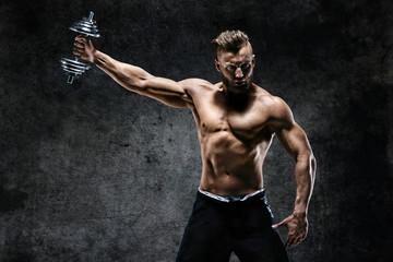 Young male bodybuilder doing exercise with heavy weight dumbbells. Photo of muscular man on dark...