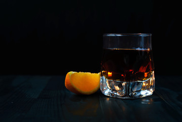 A glass of whiskey with an orange slice on a black background and a wooden table