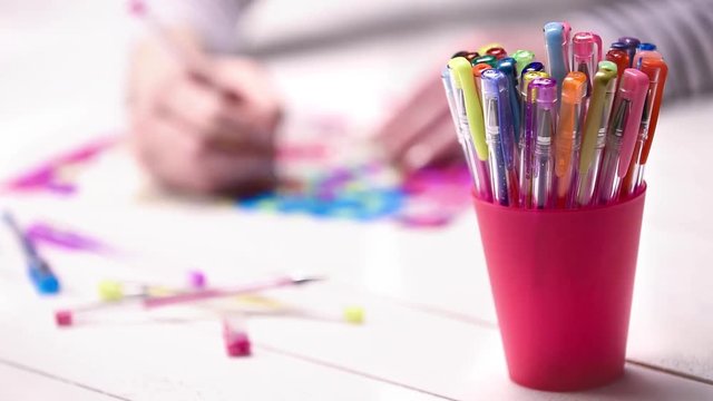 Colour footage of a young unrecognisable woman colouring in an adult colouring book, with cup full of various gel pens in the foreground. New art therapy and stress management trend.