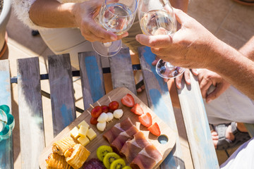 two hands senior close up drinking wine with some fruits on the table and ham and cheese