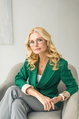 serious middle aged businesswoman in eyeglasses looking at camera while sitting in armchair
