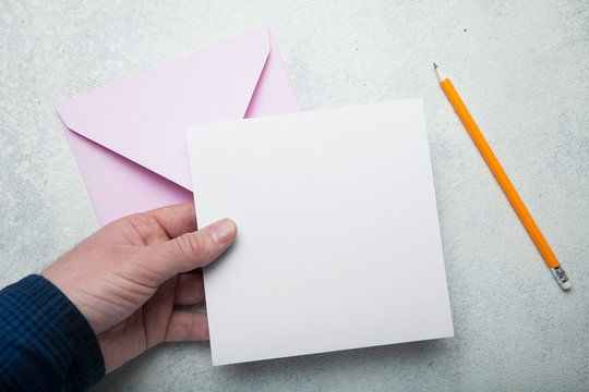 A square sheet of blank white paper in his hand, a pink envelope for mail and a yellow pencil.