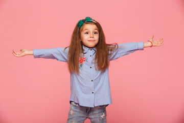 Lovely child girl in hair hoop and casual clothes giving huge hug with open hands or showing big size while being isolated against pink wall