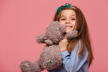 Happy girl 5-6 years having long auburn hair smiling playing with her lovely toy teddy bear isolated over pink background