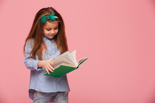 Image of smart schoolgirl 5-6 years with long auburn hair reading interesting book isolated over pink background