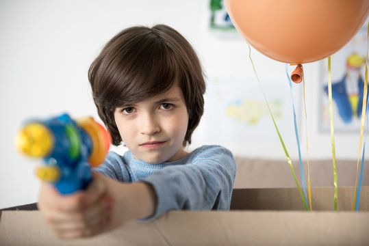 Freeze. Portrait Of Playful Boy In Cardboard Box, Kid Is Holding Water Gun With Cruel Smile. Copy Space In Right Side