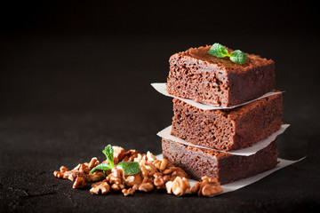 Chocolate brownie square pieces in stack on white plate with walnuts, decorated with mint leaves and cocoa on black background. Delicious dessert. Dark mood. Close up photography. Selective focus