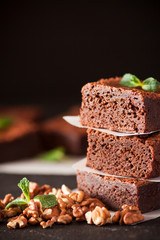 Chocolate brownie square pieces in stack on white plate with walnuts, decorated with mint leaves and cocoa on black background. Delicious dessert. Dark mood. Close up photography. Selective focus