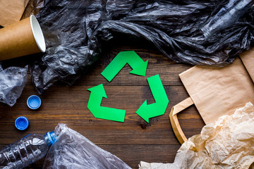 Green paper recycling sign among waste materials paper, plastic, polyethylene on dark wooden background top view close up