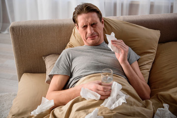 Waist up portrait of exhausted guy sneezing. He is feeling bad and having rest in bedroom while...