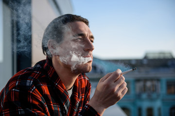 Calm adult man puffing on a cigarette with thoughtful look, he is staying on terrace. Focus on his...