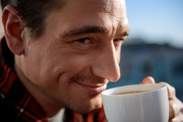 Close up portrait of contented guy looking at camera with smile, he is having a cup of drink in his hands. Focus on his face