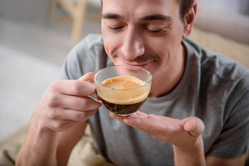 Close up portrait of peaceful guy smelling drink and smiling, he is sitting on bed and holding a cup