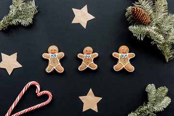 Three gigngerbread men on a black backdrop surrounded by papr stars, small fir tree 	branches, a cone and two candy canes