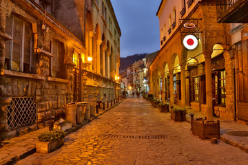 Fototapeta premium Illuminated cobbled street with light reflections on cobblestones in old historical city by night. Dark blurred silhouette of person evokes Jack the Ripper.