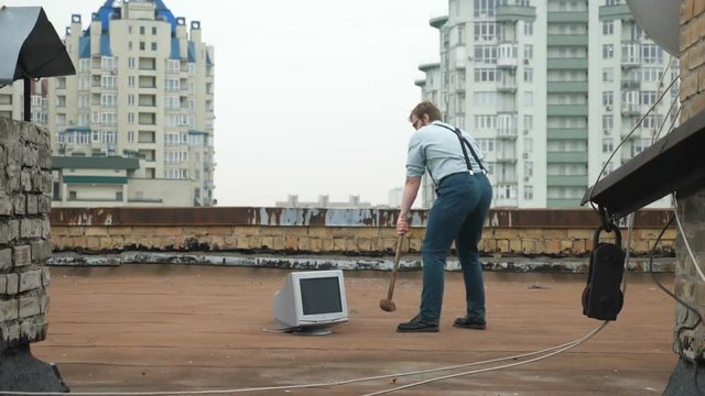 Young strong man smashes the monitor with a sledgehammer on the roof. Hammer, bat, violence, hatred, anarchy, destruction. 60 fps