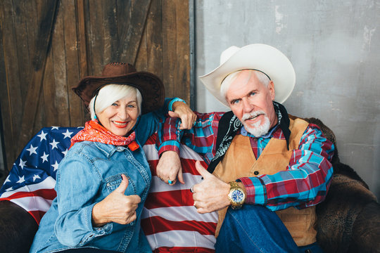 Elderly couple dressed in cowboy hats, showing thumbs up, smiling and looking at camera. Behind, on the couch lies the American flag, the celebration of America's Independence Day