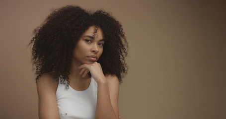 mixed race black woman portrait with big afro hair, curly hair in beige background. Thinking about something with a hand on chin