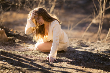 Young mixed race woman in white dress crawls on ground by trees destroyed by wildfire while covered...