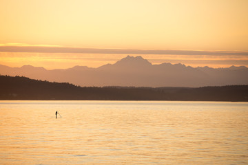 Silhouetted stand up paddle board rider on a calm ocean, with mountains on the horizon and orange sunset