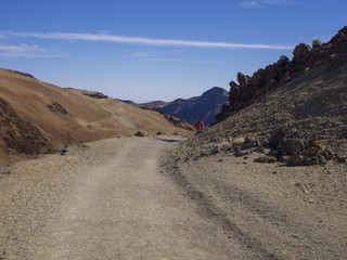desert volcanic landscape with couple of hikers in red clothes walking on road to volcano pico del teide with orange and purple mountain with clear blue sky background