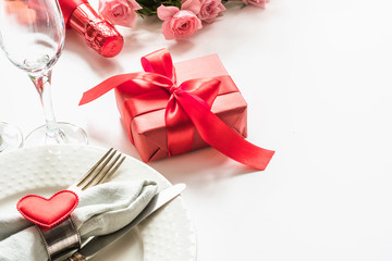 Valentines day dinner with table place setting with red gift, glass for champagne, a bottle of champagne, heart ornaments with silverware on white. Close up. Valentine's card.