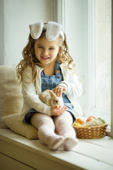 Cute happy little child girl is wearing bunny ears on Easter day sitting on window sill holding her friend little colorful rabbit