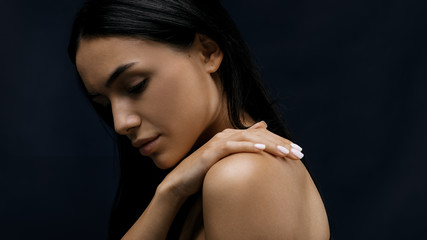Fototapeta premium Side view of a beautiful woman with black hair touching her shoulder with hand against black background