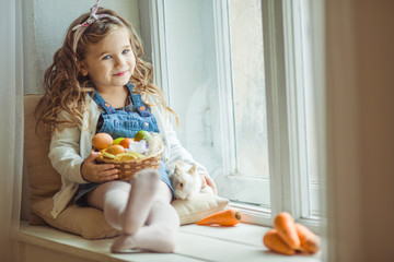 Beautiful happy child girl is sitting on window sill with her friend rabbit and holding basket with painted eggs, Easter holiday concept