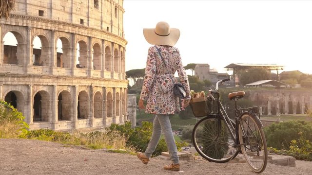 Young woman tourist with bycicle takes pictures of the colosseum in rome at sunset with smartphone. Beautiful stylish dress with large hat, flowers and bread in basket.