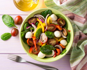 Fresh salad with spinach, bell peppers, tomatoes, red onions and mozzarella on purple wooden background.