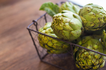 Fresh organic artichokes over a dark blue napkin with knife and lemon and with metal basket with artichokes over a rustic wooden background. Top View