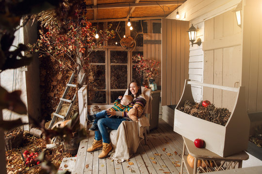 Cute Little Baby Boy And Mother Dressed In A Sweater, Jeans Playing Near House With Plush Toy Teddy Bear In Autumn Time. Woman And Son On Courtyard, Lit By Flashlights, With Dry Fall Leaves, Pumpkins.