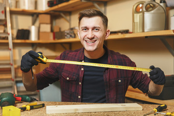 Handsome caucasian young man in plaid shirt, black T-shirt, gloves measuring length of piece of wood with tape measure, working in carpentry workshop at wooden table place with different work tools.
