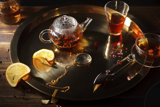 Small glass teapot with hot black tea, cup, glasses, dried rose petals, pocket magnifier on golden chain, squeezed orange slice on golden tray on wooden background. Evening light. Overhead view.