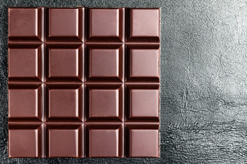 Dark, bitter chocolate bar on black leather texture background, top view