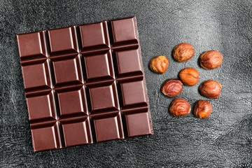 Dark, bitter chocolate bar and hazelnuts on black leather texture background, top view