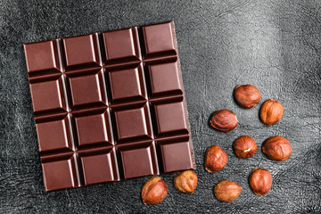 Dark, bitter chocolate bar and hazelnuts on black leather texture background, top view