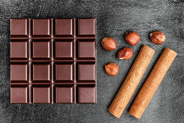 Dark, bitter chocolate bar, hazelnuts and two cinnamon sticks on black leather texture background, top view