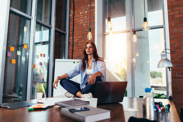 Young businesswoman meditating sitting on working table in lotus pose in her office