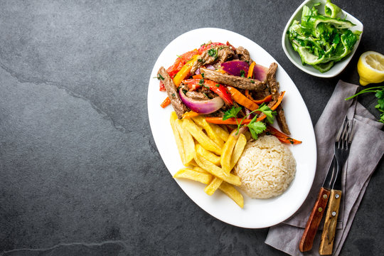 Peruvian dish Lomo saltado - beef tenderloin with purple onion, yellow chili, tomatoes served on white plate, slate background