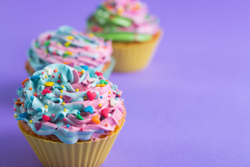 Three cupcakes with creamy multicolored top with colorful hearts and sprinkles on violet background